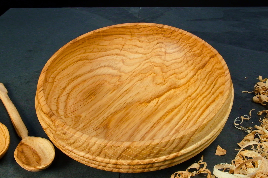 A wooden bowl on a table next to some wood shavings and hand-carved wood spoons
